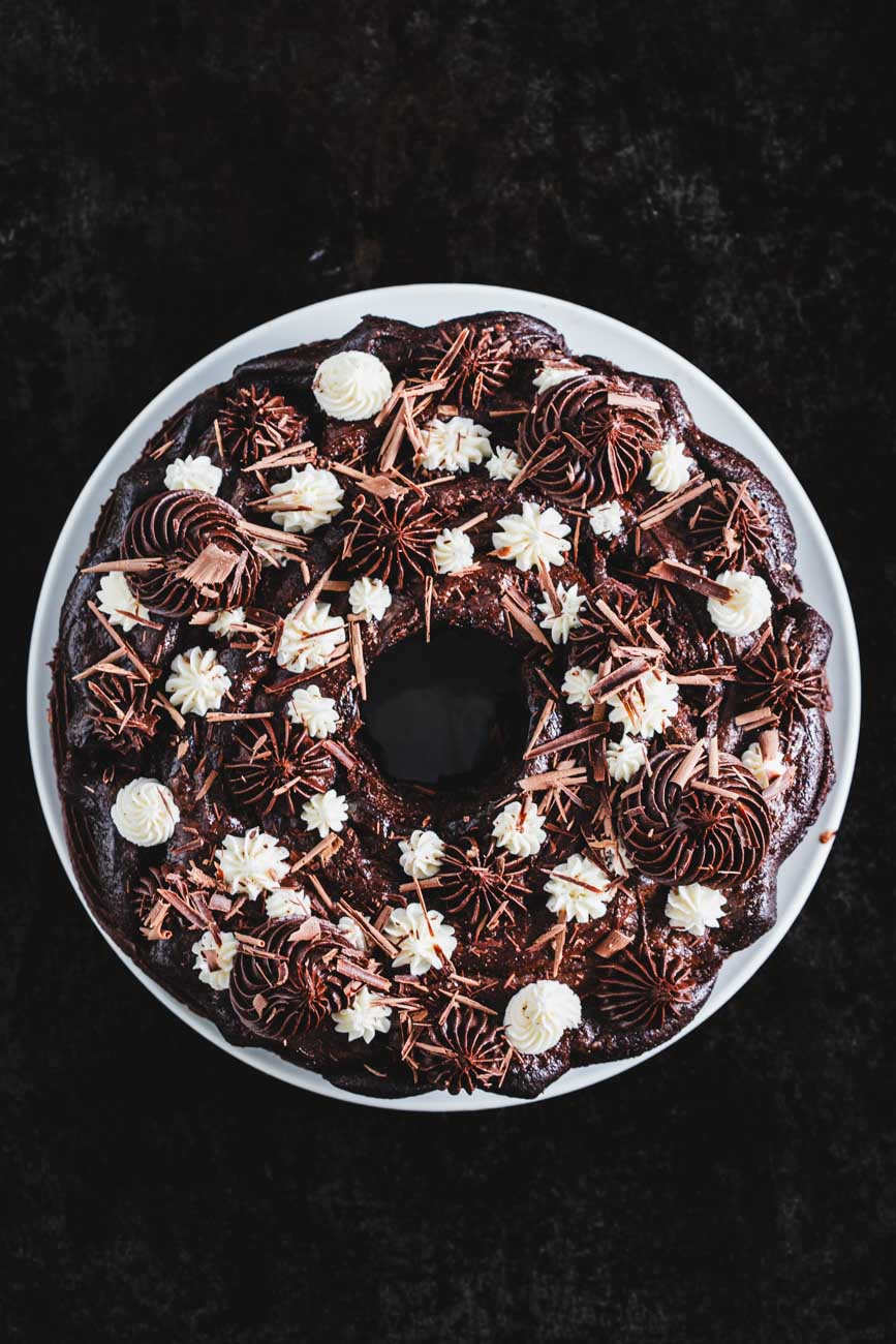 Top view of a cocoa bean bundt cake decorated with chocolate and vanilla frosting swirls and chocolate shavings on a white plate.
