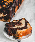A slice of marble loaf cake on a white plate, showcasing the intricate swirl pattern of chocolate and vanilla, with the rest of the loaf visible in the background.