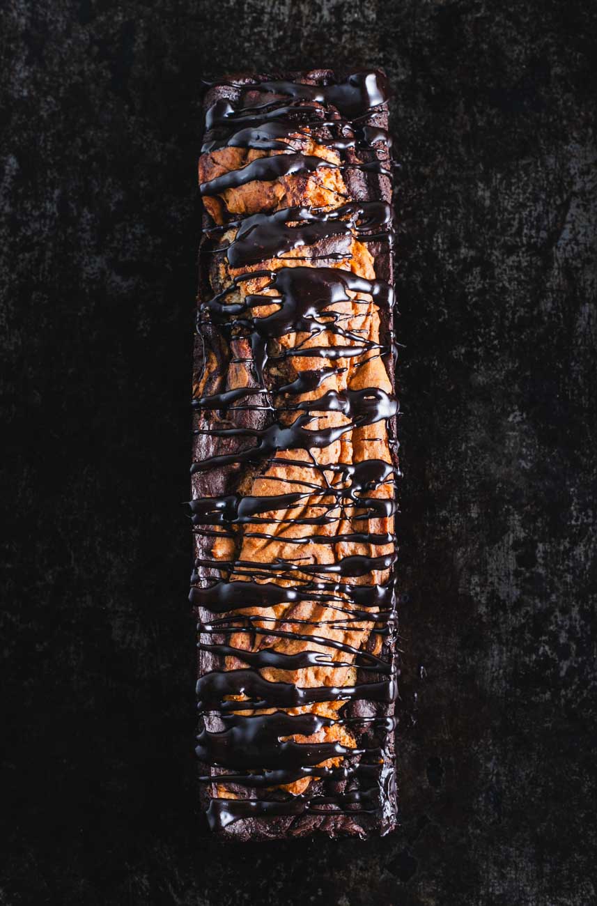  A top-down view of a whole marble loaf cake, drizzled with dark chocolate ganache on a dark surface.