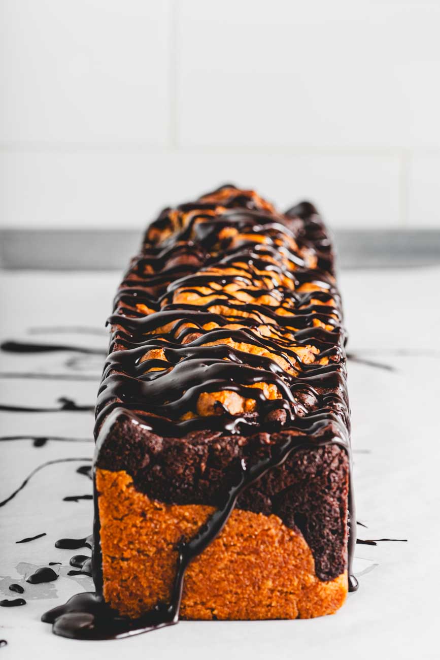 A side view of a whole marble loaf cake on a white surface, with chocolate ganache dripping down the sides.