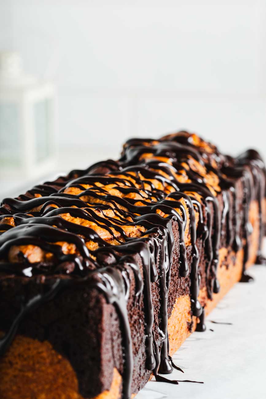 A side-angle view of a whole marble loaf cake, with a thick drizzle of dark chocolate ganache.