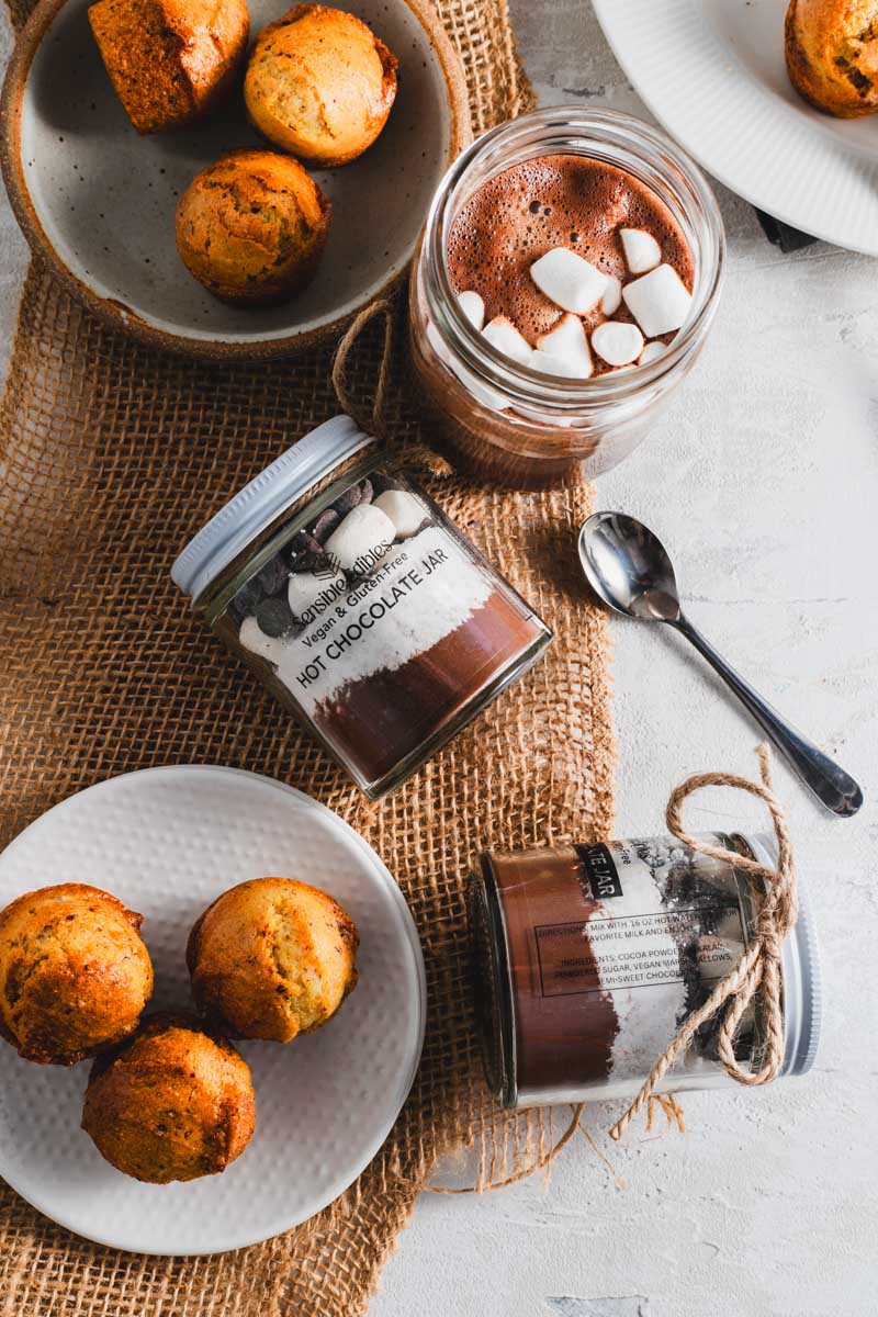 Hot chocolate jar with marshmallows, muffins, and a cup of hot chocolate on a burlap cloth.