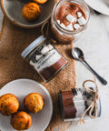 Hot chocolate jar with marshmallows, muffins, and a cup of hot chocolate on a burlap cloth.