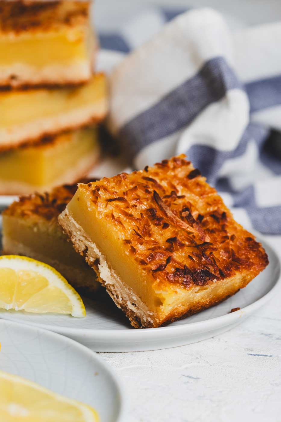 close up of a lemon bar with toasted coconut topping on a plate with a stack of lemon bars in the background and lemon slices. 