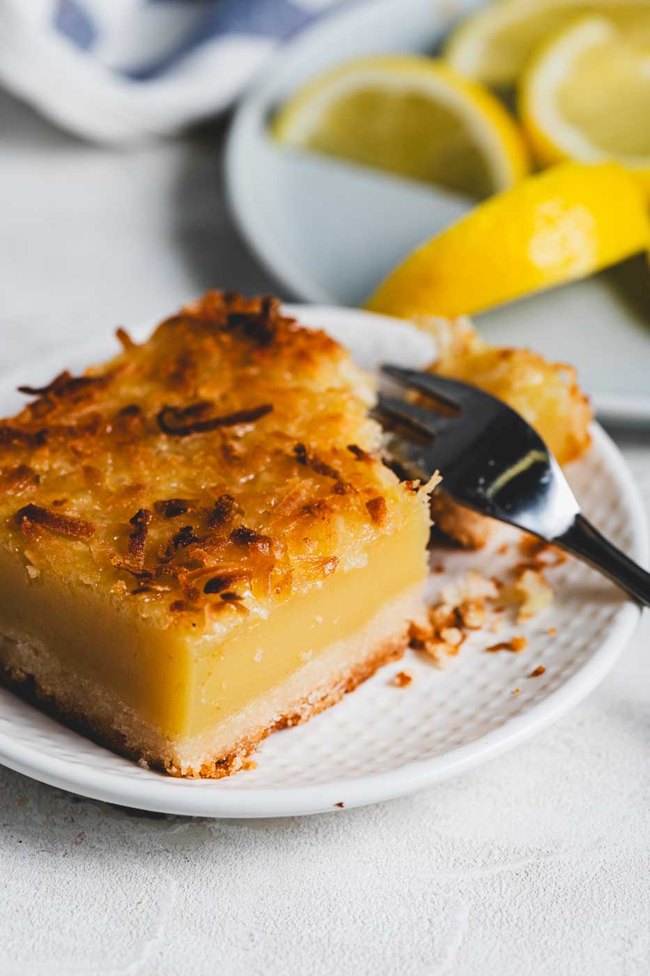 close up of a lemon bar with toasted coconut topping on a plate with a fork breaking it apart. There are slices of lemon in the background.