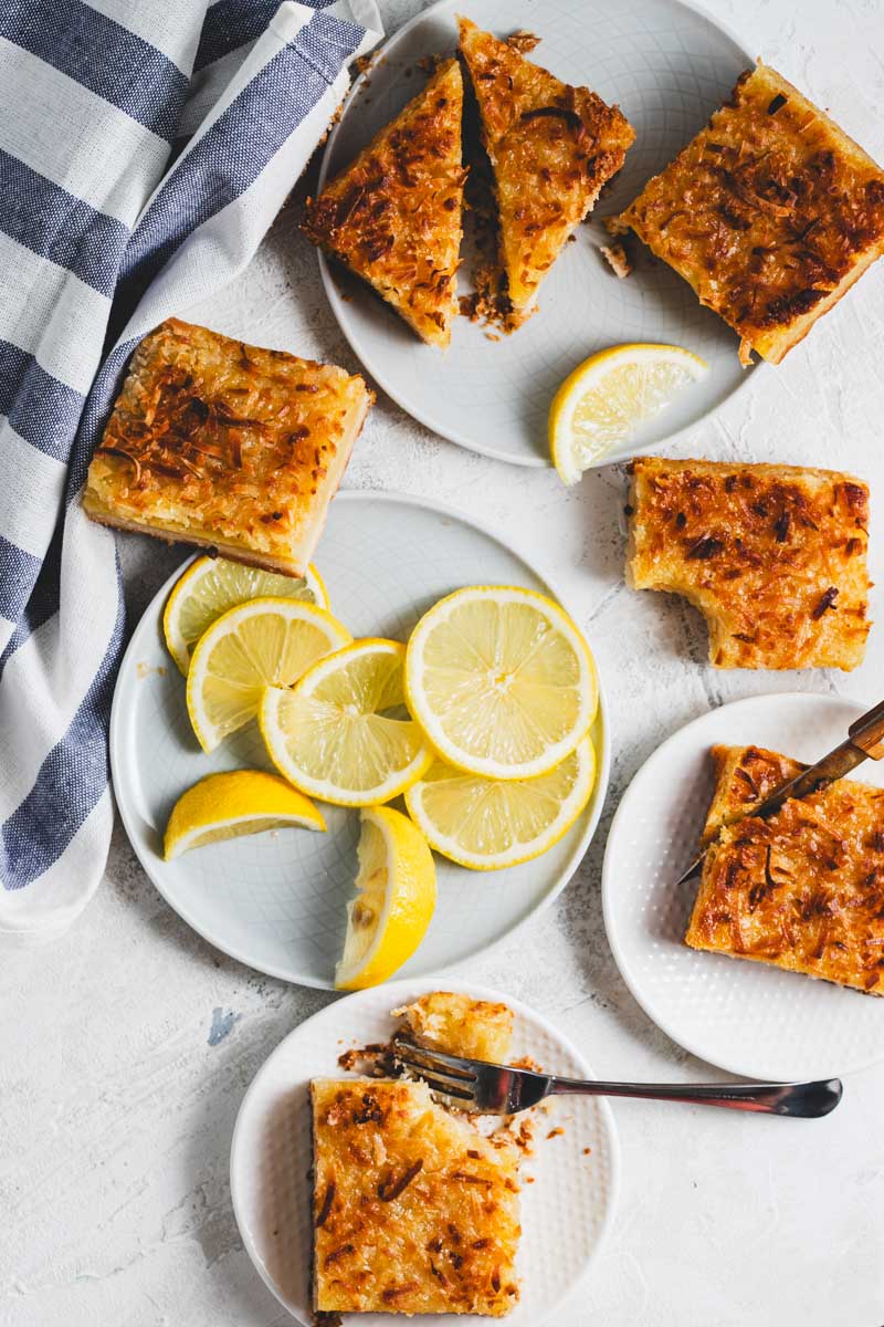 Lemon bars with golden-brown toasted coconut shavings on top arranged across several white plates with slices of lemon, some being cut apart with a knife and fork
