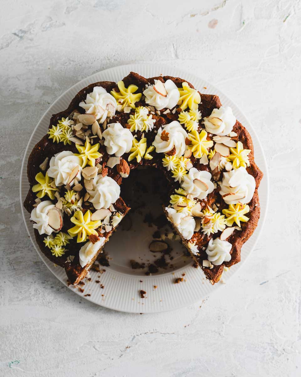 Top view of a lemon almond bundt cake with a slice removed, exposing the soft, moist interior. The cake is decorated with dollops of white and yellow frosting and almond slices.