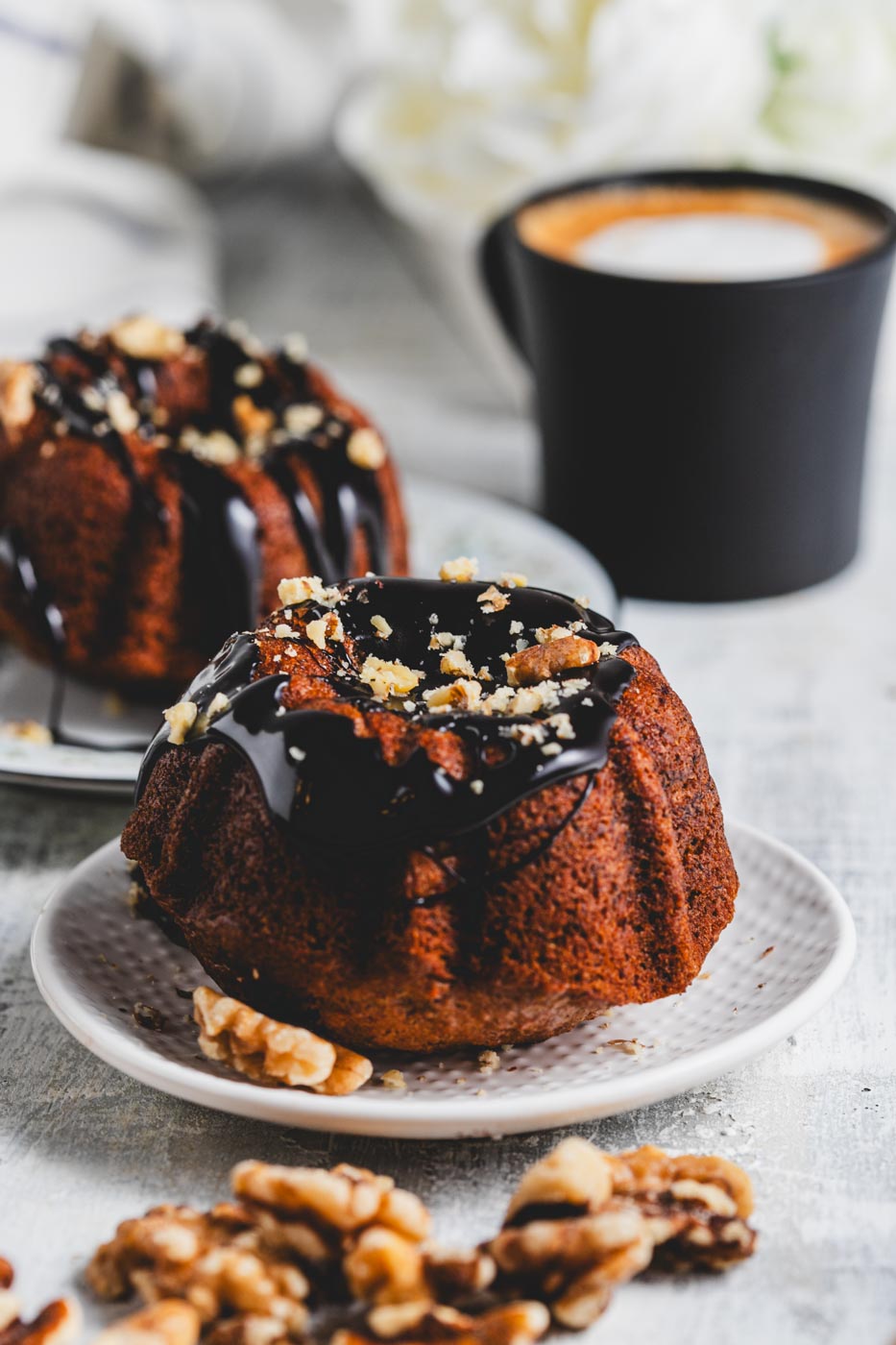 Mini banana bundt cakes with chocolate glaze and crushed walnuts on white plates with a mug of coffee and whole walnuts scattered around