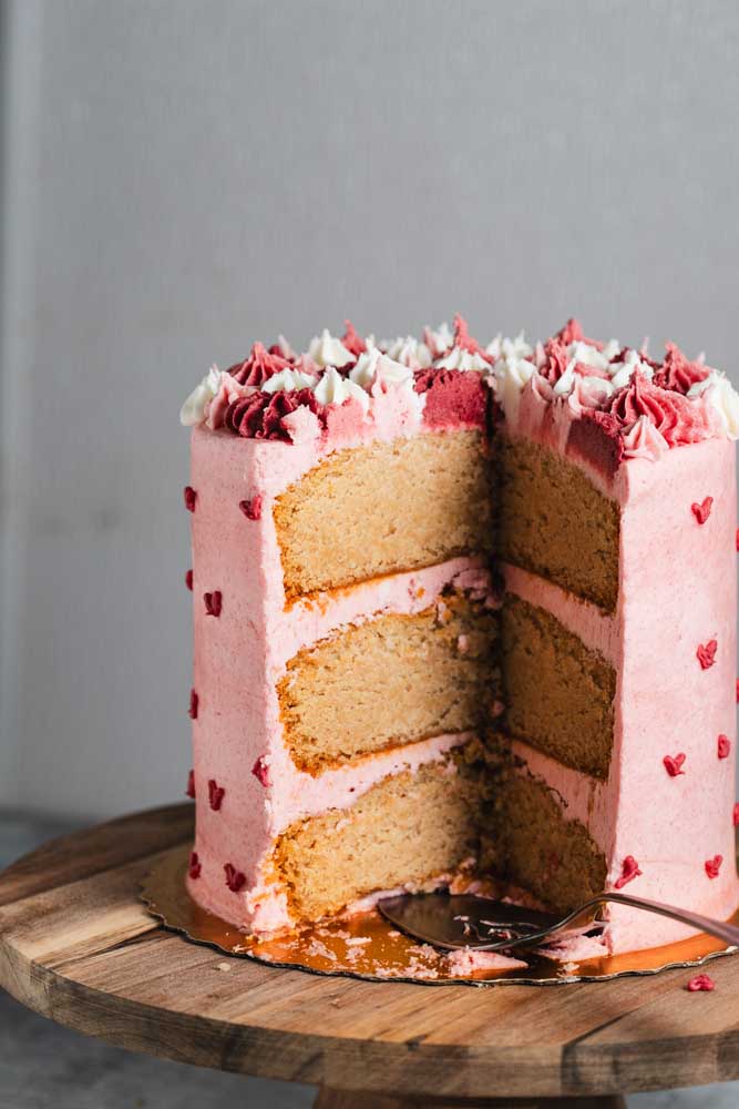 A layered pink velvet cake with pink frosting with decorative pink hearts and pink and white frosting decorations with a slice taken out