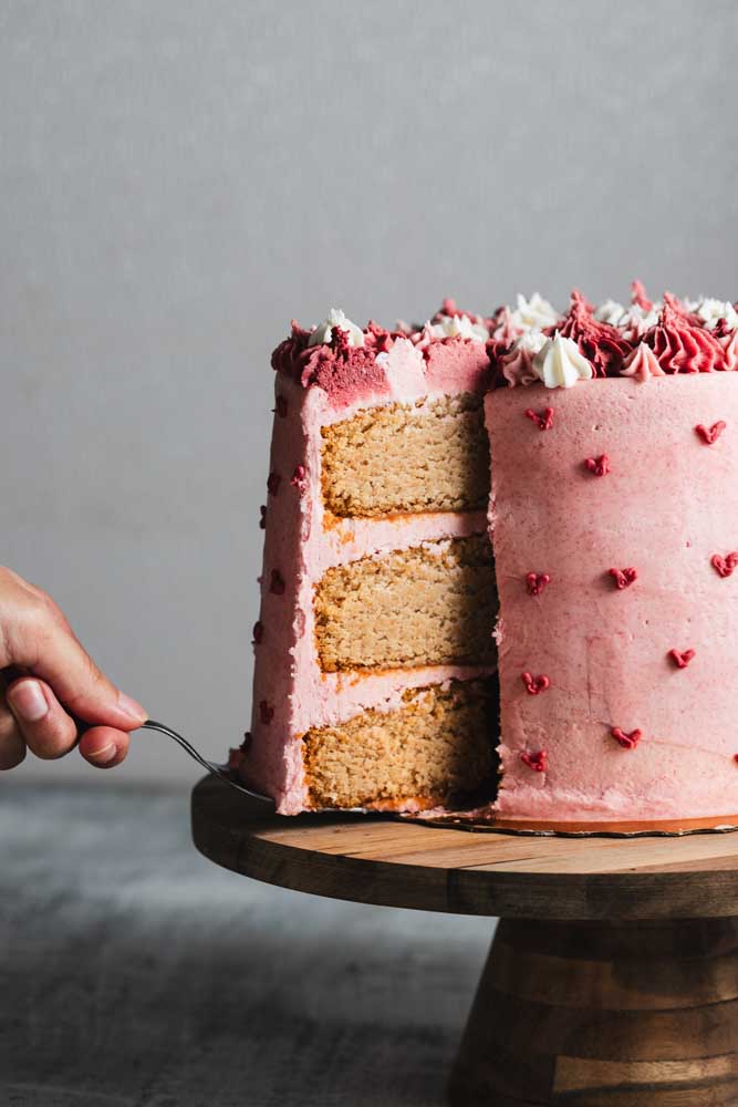 A layered pink velvet cake with pink frosting with decorative pink hearts and pink and white frosting decorations with a slice being taken out