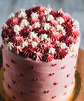 A layered pink velvet cake with white and red frosting decorations, placed on a wooden stand.