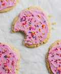 A set of pink frosted sugar cookies with rainbow sprinkles on top, and one half-eaten cookie showing the creamy pink icing and sprinkles.