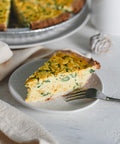 A slice of vegan quiche on a white plate with a fork and the rest of the quiche in the background