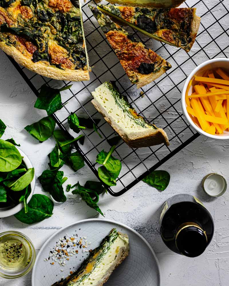 Slices of spinach cheddar quiche on a cooling rack with spinach leaves scatted and a bowl of cheddar.
