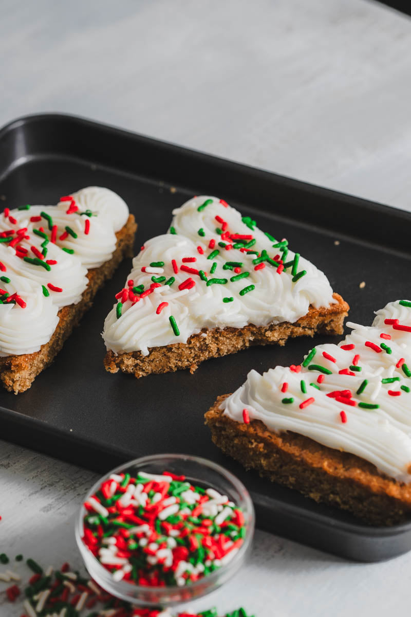 Christmas tree-shaped cookies with white frosting and red and green sprinkles on a black tray, with a bowl of sprinkles.