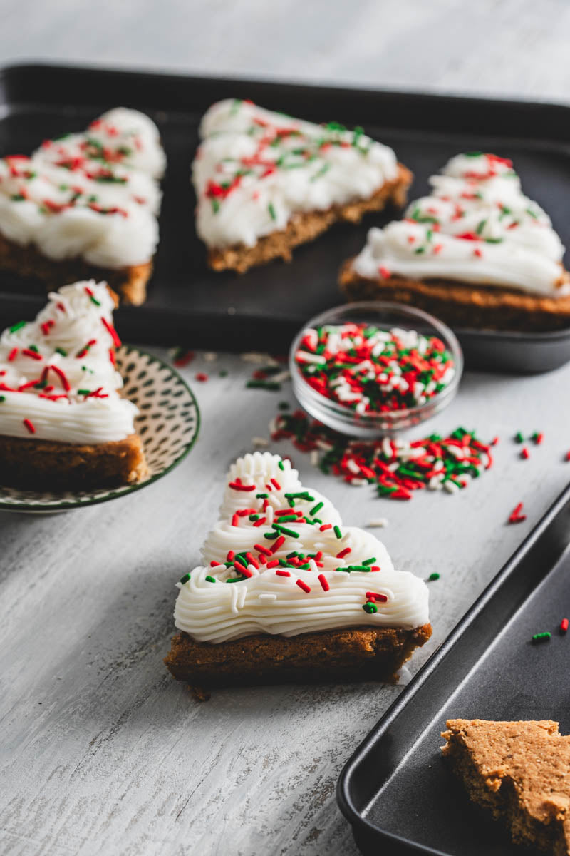 Close-up of Christmas tree-shaped cookies with white frosting and red and green sprinkles on a black tray, with a bowl of sprinkles.