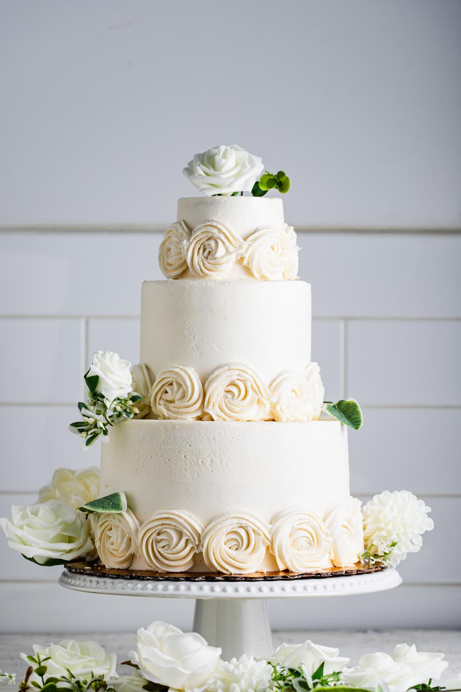 An elegant three tiered wedding cake with white frosting and white swirled frosting flowers around the base of each tier. There are decorative white flowers around and on top of the cake.