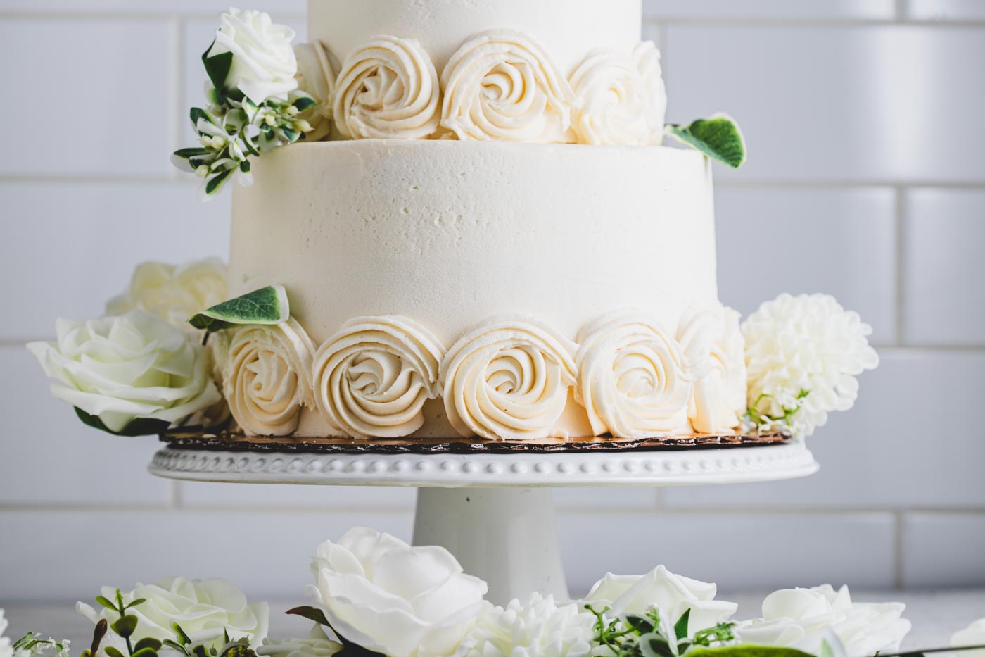 A close up of a tiered wedding cake on a white cake stand showing detail of white frosting flowers and decorative white flowers