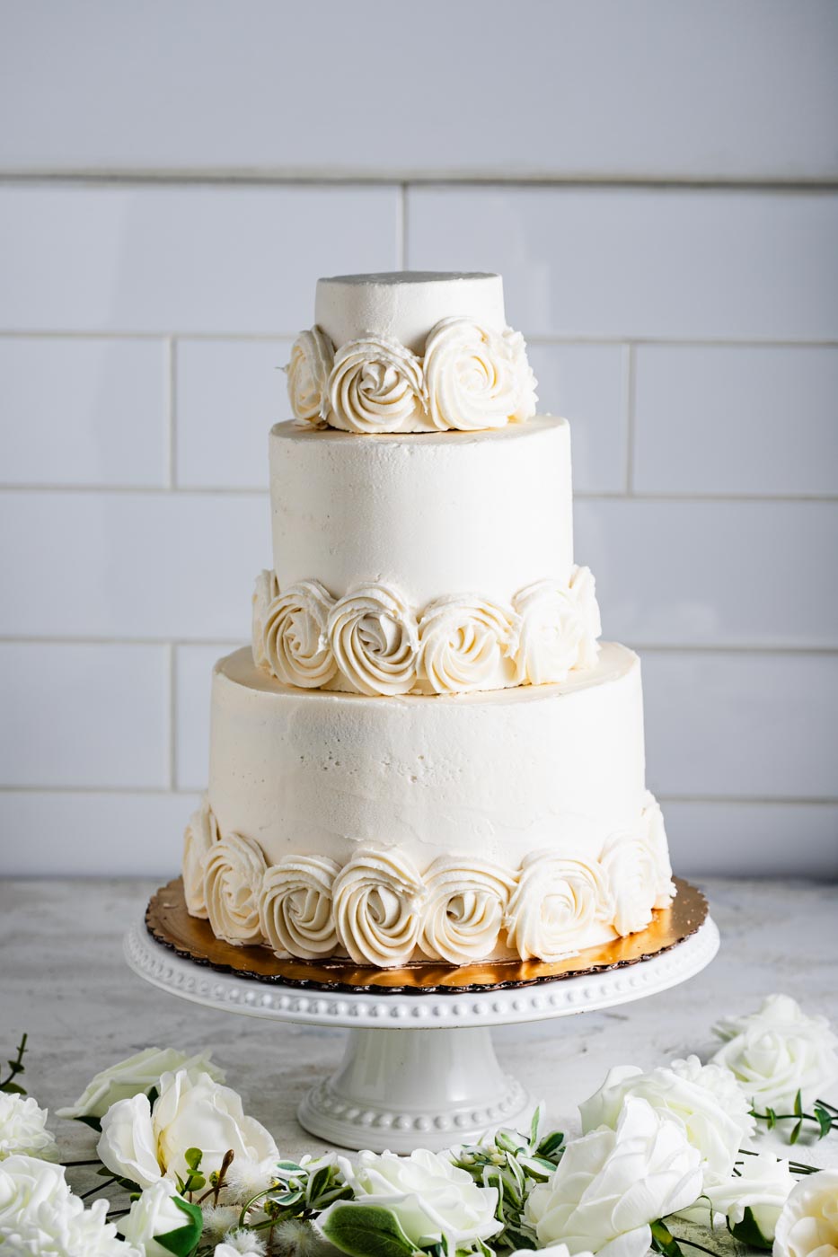An elegant three tiered wedding cake with white frosting and white swirled frosting flowers around the base of each tier. There are decorative white flowers around the cake.
