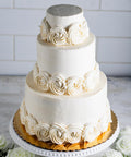 An elegant three tiered wedding cake with white frosting and white swirled frosting flowers around the base of each tier. There are decorative white flowers around the cake.