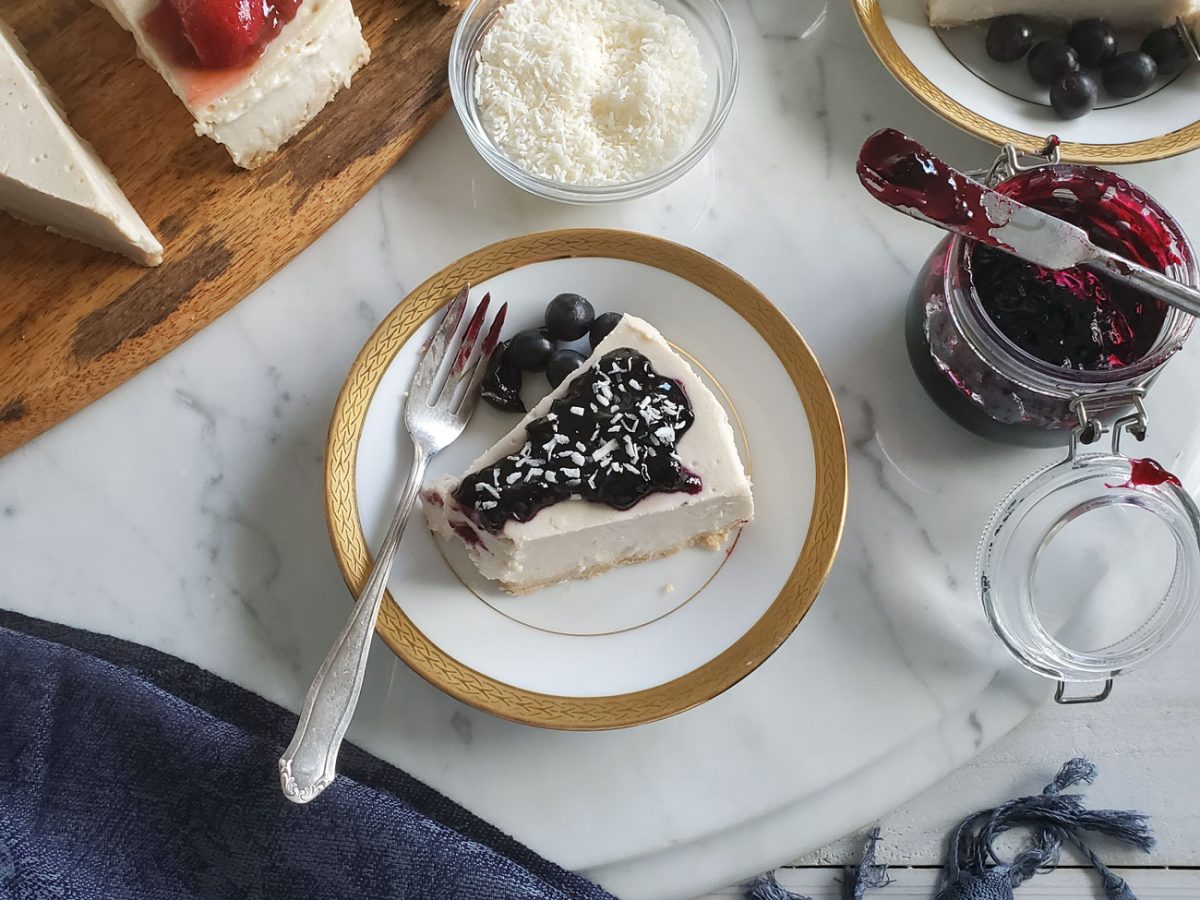 Slice of vegan cheesecake topped with blueberry compote on a white plate with a gold rim, accompanied by a jar of compote and a fork, set on a marble surface.