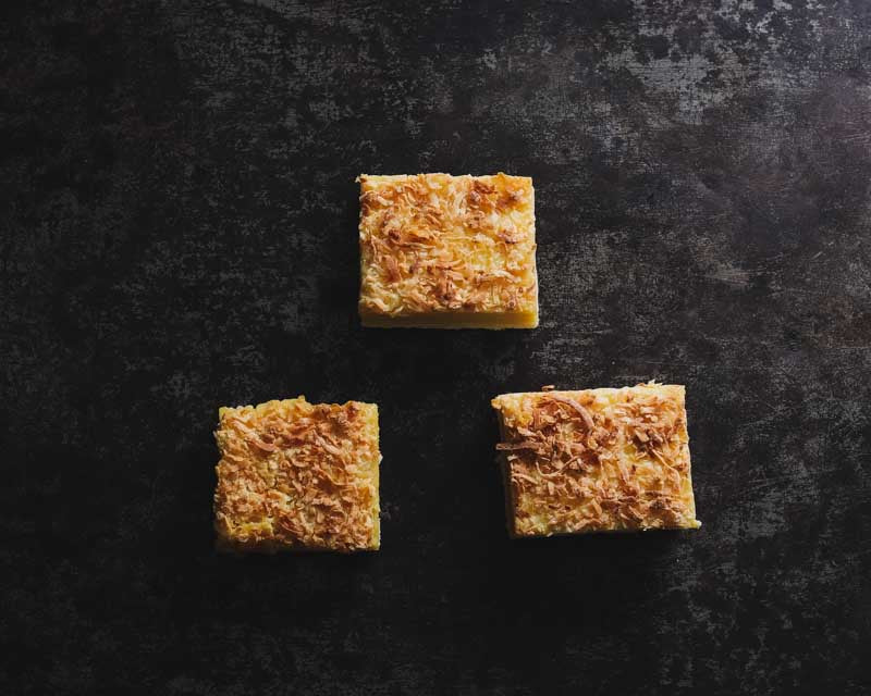 Three square lemon bars topped with coconut shavings on a dark surface.