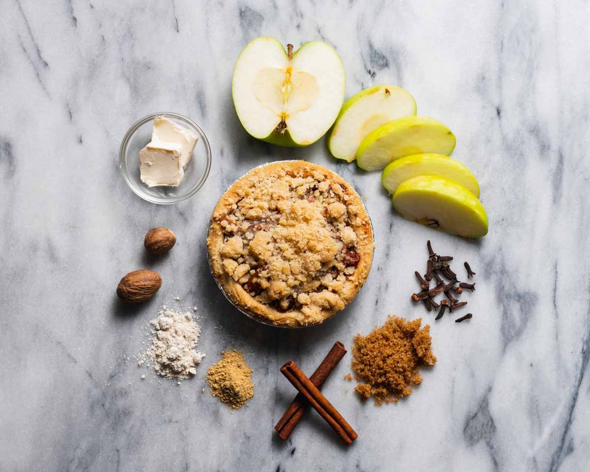 Mini apple crumb pie on a marble surface, surrounded by apple slices, cinnamon sticks, and brown sugar.