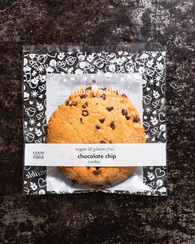 Packaged chocolate chip cookie from Sensible Edibles, wrapped in clear plastic with a white label, displayed on a dark rustic surface.
