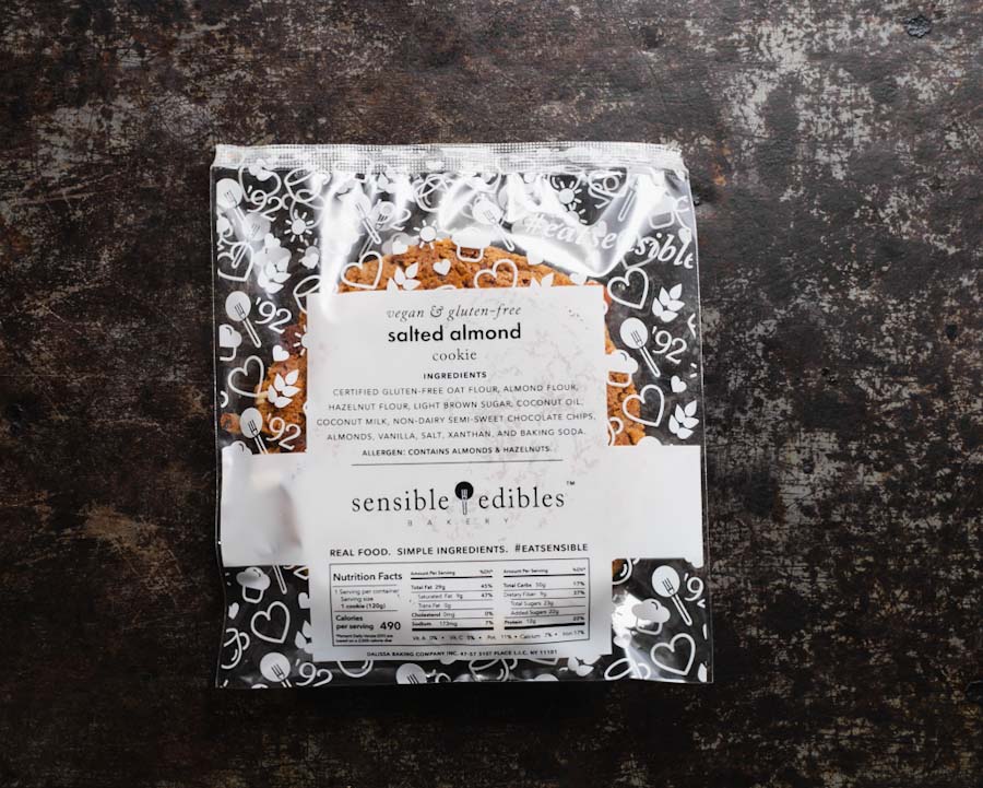 Back view of packaged salted almond chocolate chip cookie from Sensible Edibles Bakery showing ingredients and nutritional facts on a dark rustic background.