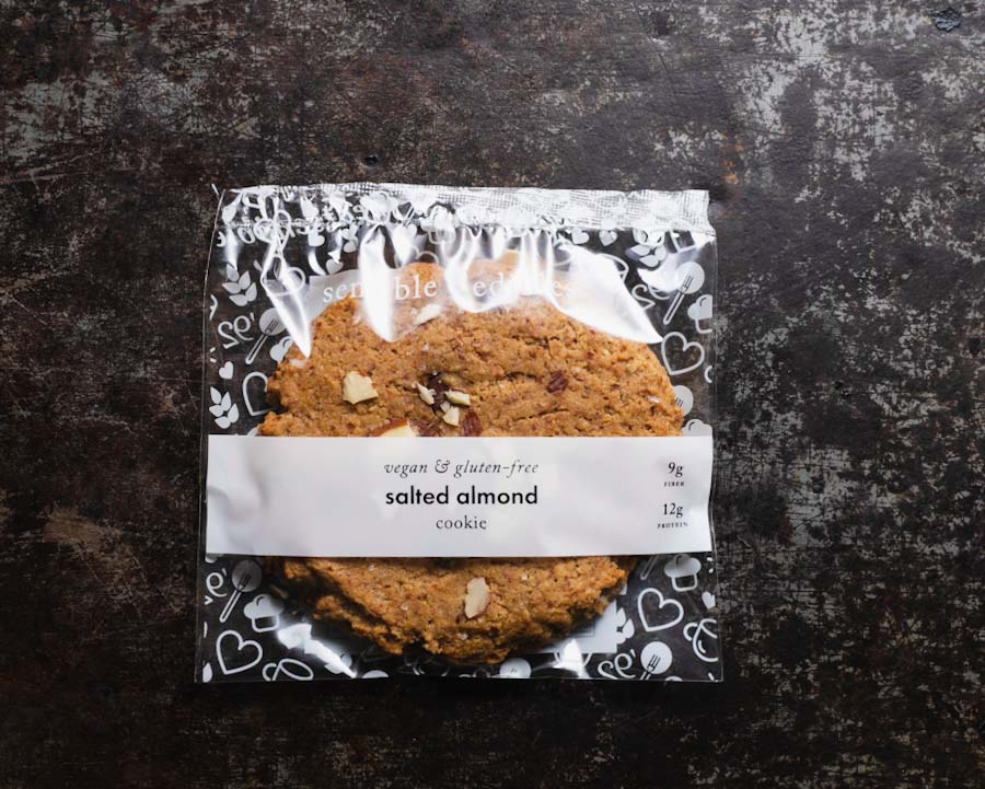 Packaged salted almond chocolate chip cookie from Sensible Edibles Bakery with clear floral packaging showing the label on a dark rustic background.