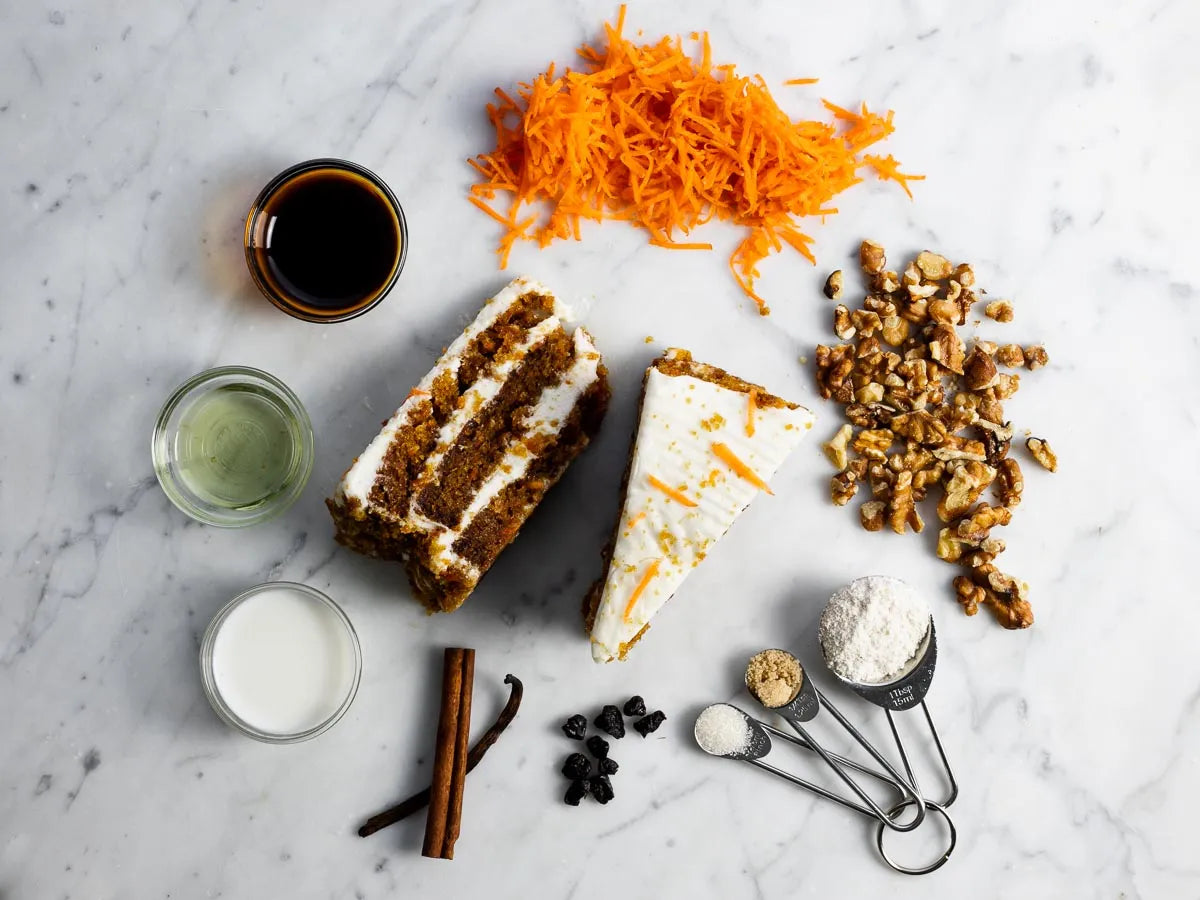 Ingredients for carrot cake, including grated carrots, walnuts, flour, cinnamon sticks, raisins, milk, oil, vanilla, baking powder, and a slice of frosted carrot cake, arranged on a marble surface.