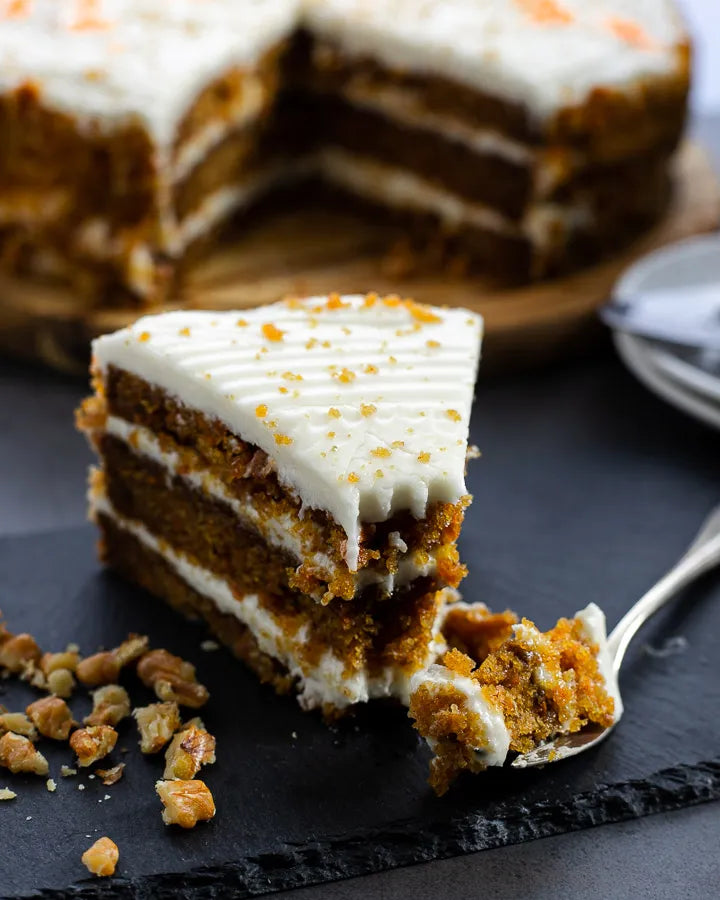 Slice of carrot cake with creamy frosting layers on a slate board, topped with crushed walnuts.