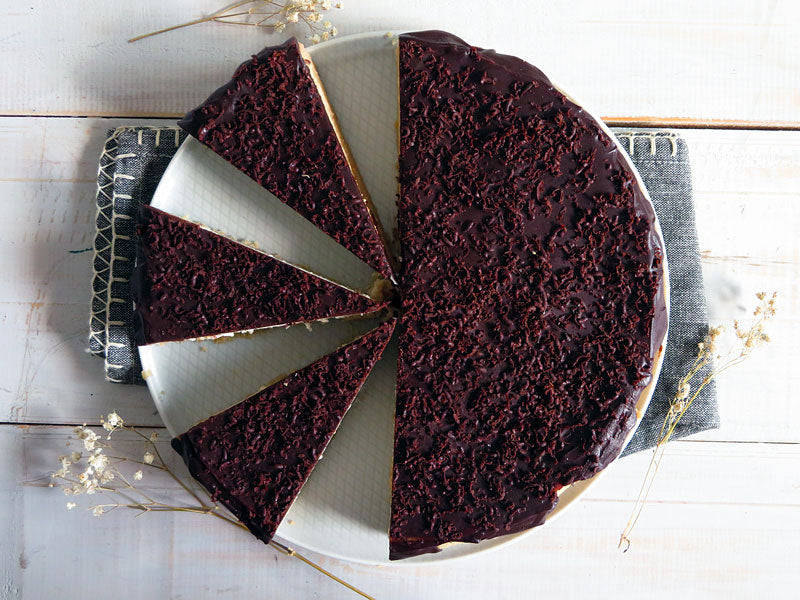 Top view of a chocolate cheesecake with a chocolate topping, sliced into pieces on a white plate, placed on a grey cloth with dried flowers.