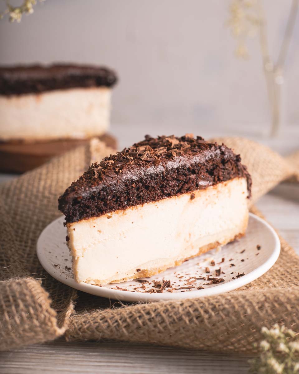 Slice of chocolate cheesecake with a creamy filling and chocolate topping, served on a white plate with chocolate shavings, placed on a rustic burlap cloth.