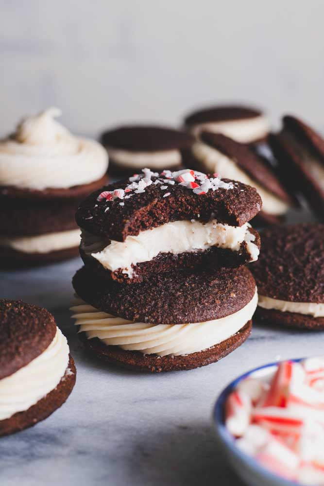 Stack of chocolate whoopie pies with peppermint cream filling topped with crushed peppermint candy, one with a bite taken, showing the creamy interior, on a light surface.