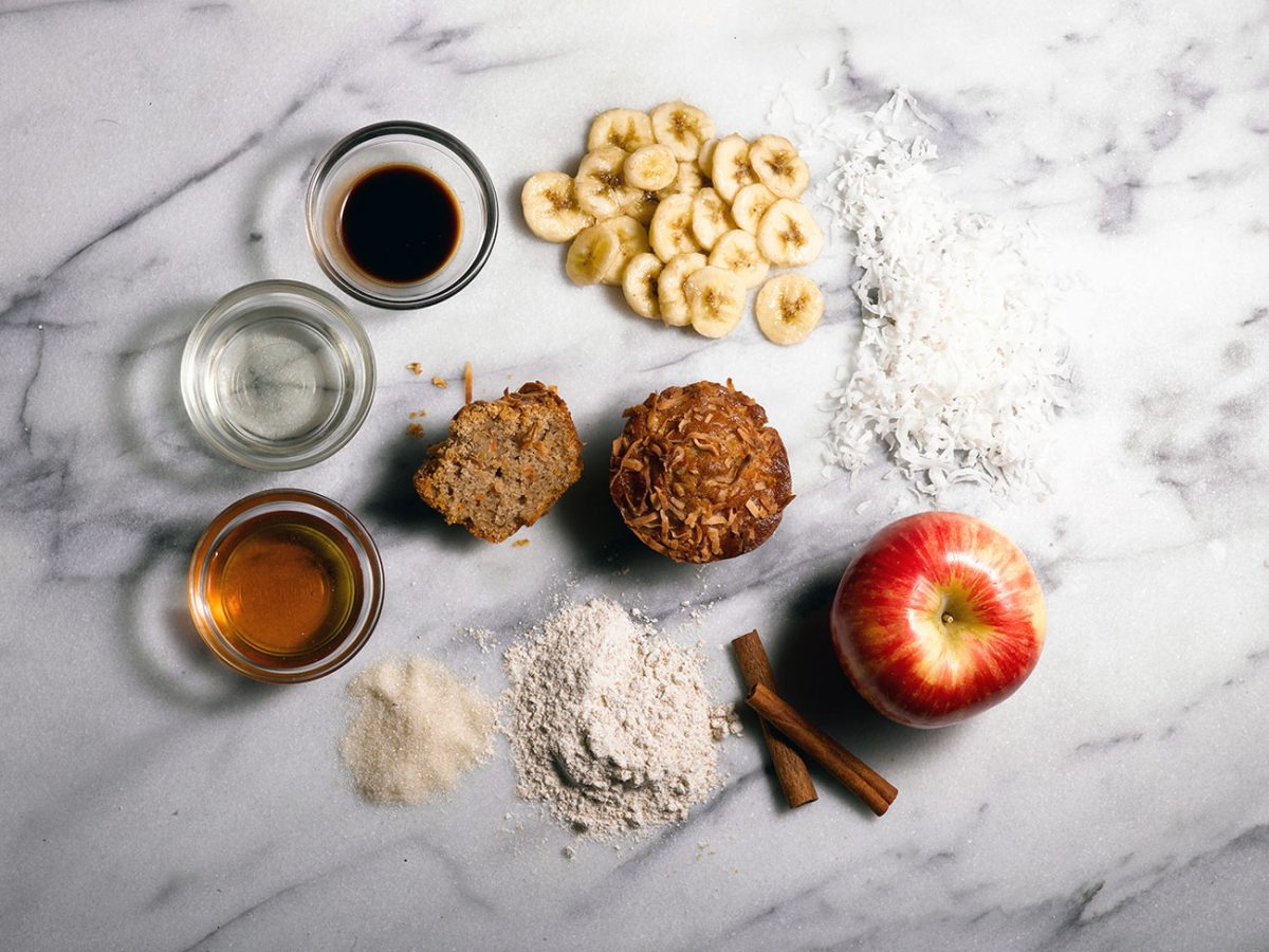 Ingredients for harvest muffin laid out on a marble surface, including banana slices, shredded coconut, apple, and cinnamon.