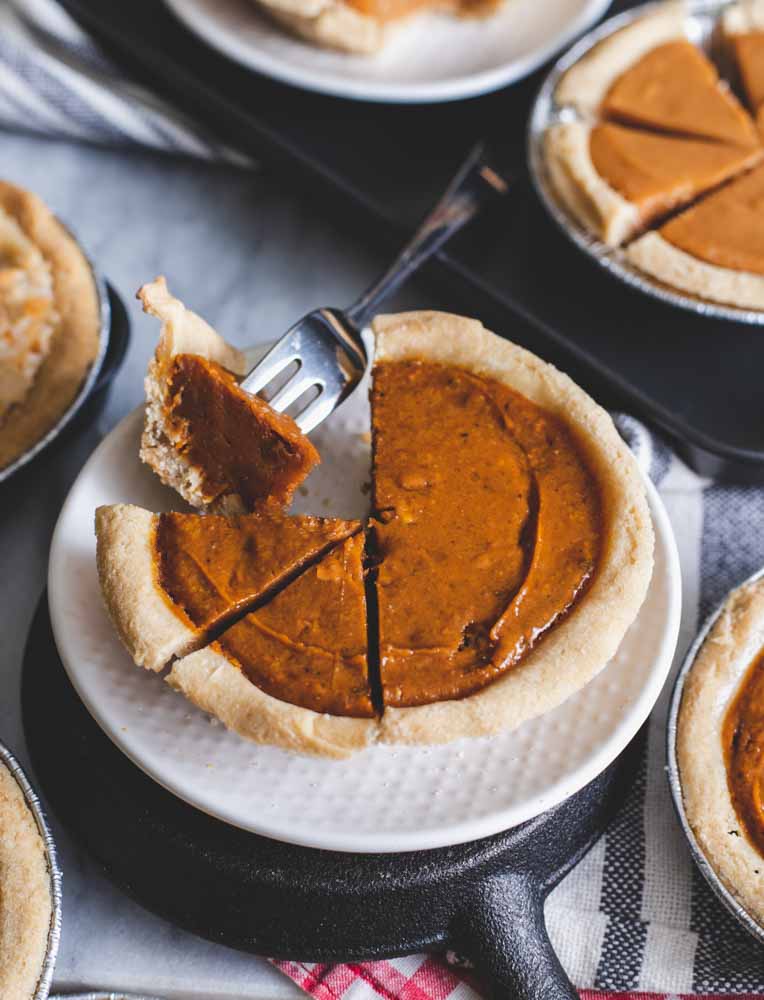 A mini pumpkin pie with a slice cut out, placed on a white plate, showing a fork holding a piece of pie, with more pies in the background.