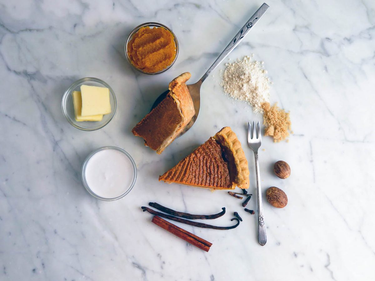 Ingredients for mini pumpkin pies including pumpkin puree, butter, coconut cream, flour, brown sugar, nutmeg, vanilla bean, and cinnamon sticks on a marble surface.