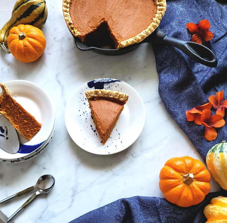 A slice of pumpkin pie on a white plate with a blue pattern, with the whole pie in a cast iron skillet and mini pumpkins on a marble surface decorated with blue cloth and orange flowers.
