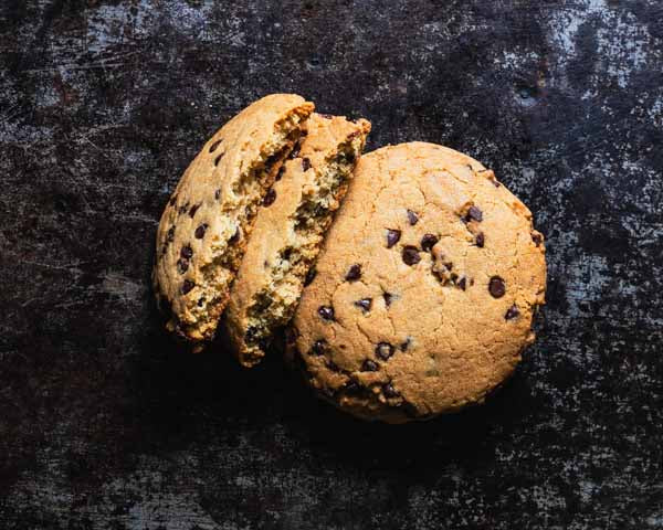 Two chocolate chip cookies on a dark rustic surface, with one cookie broken in half to reveal the inside texture.