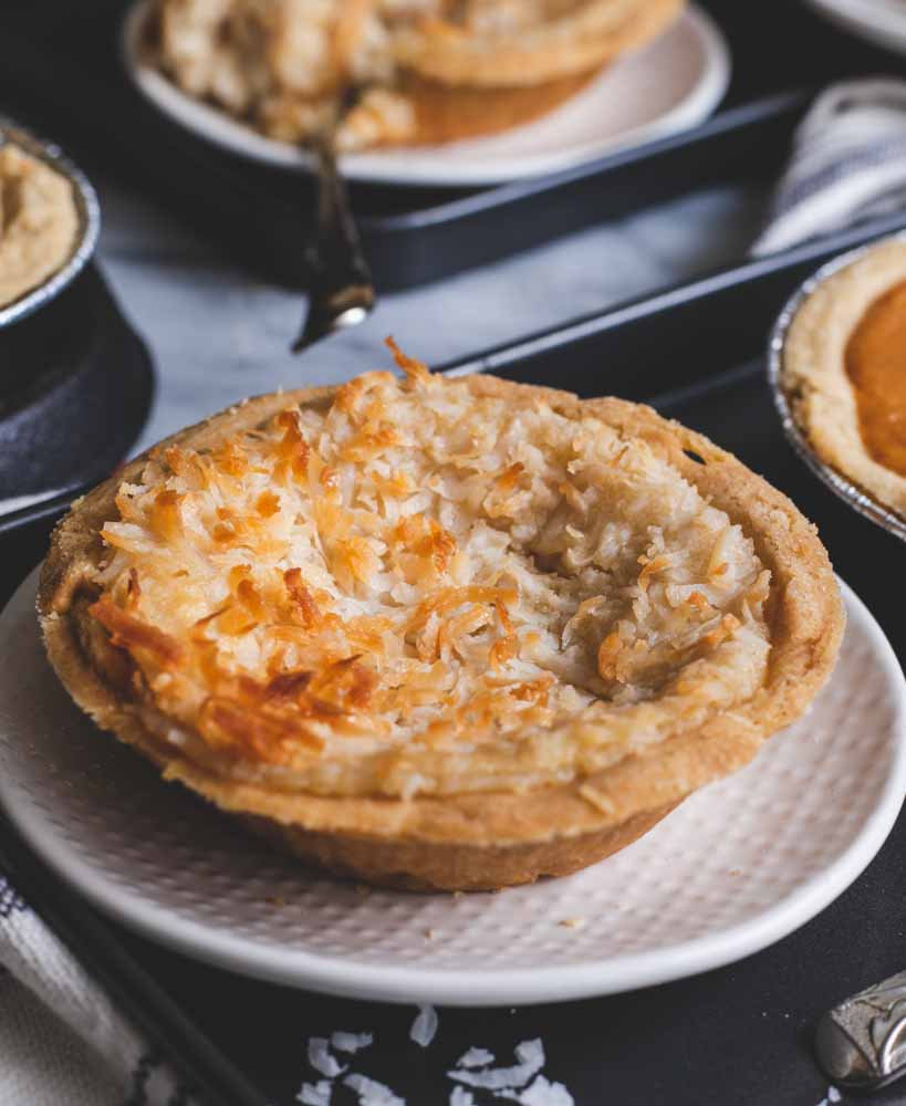 Mini coconut custard pie with toasted coconut on a white plate, surrounded by other pies on a baking tray.