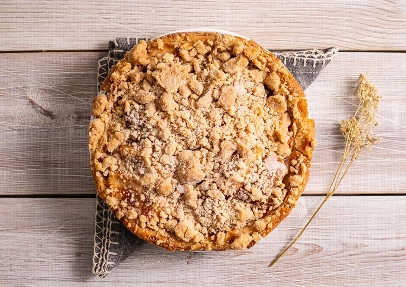 Large apple crumb pie on a rustic wooden table with decorative napkin and dried flowers