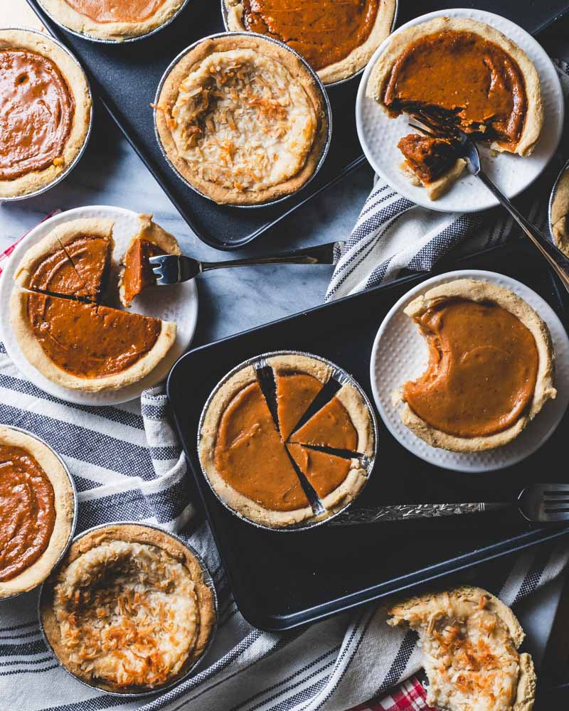  Various mini pumpkin pies, some sliced and some whole, arranged on black trays and white plates, with forks and a striped cloth on a marble surface.